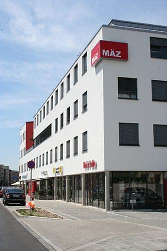 Nahansicht der Gebäudeecke mit MÄZ-Schild, Fensterreihen und verglastem Erdgeschoss an der Ostendstraße