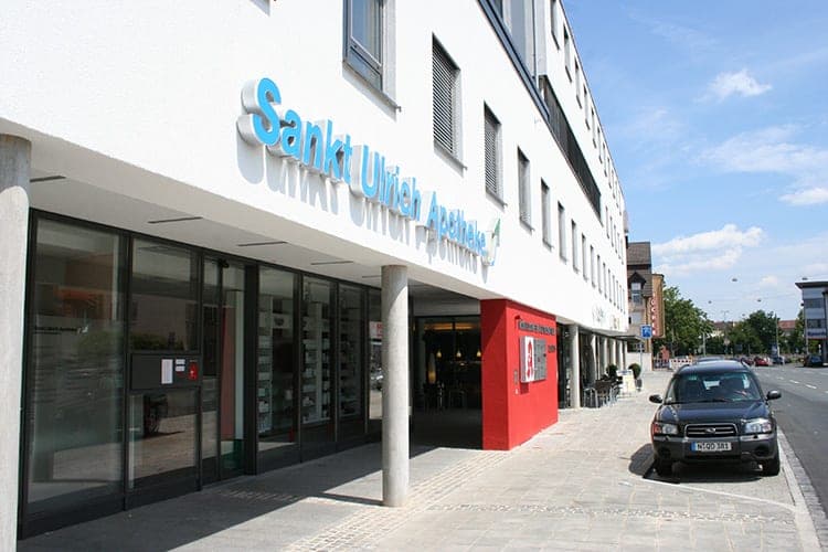 Eingang der Sankt-Ulrich-Apotheke mit Glasfront, Stützen und rotem Wandfeld unter dem auskragenden Obergeschoss