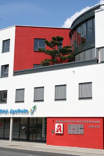 Fassadenausschnitt mit rotem Wandfeld, Kiefer auf der Dachterrasse und dem Schild des Ärztezentrums