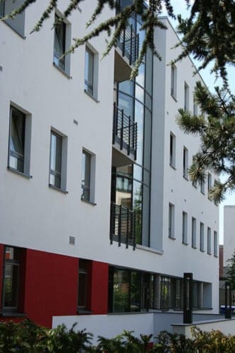Weiße Lochfassade mit verglastem Balkonband und rotem Sockel, im Vordergrund Zweige eines Nadelbaums