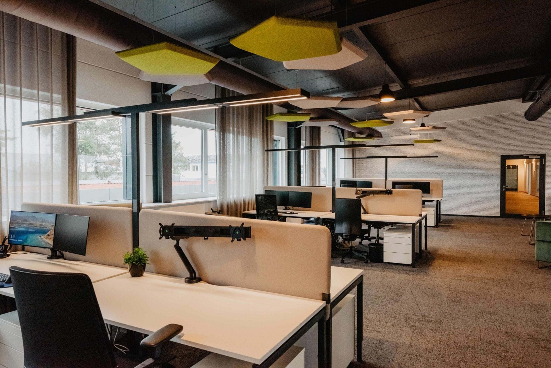 Open-plan office with desk clusters, floor lamps and coloured acoustic baffles below the dark ceiling