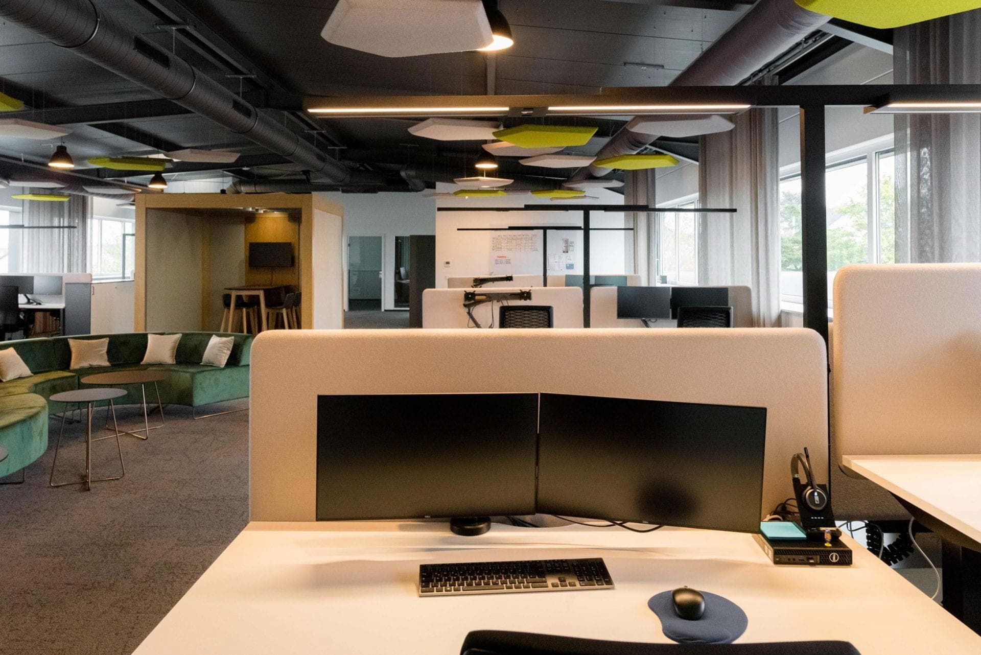 Workstation with two monitors in the foreground, with the lounge, meeting pod and further desks beyond