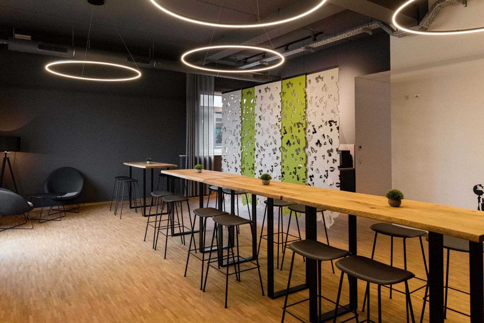 Break area with long wooden tables, bar stools, ring lights and a partition with a white and green leaf pattern