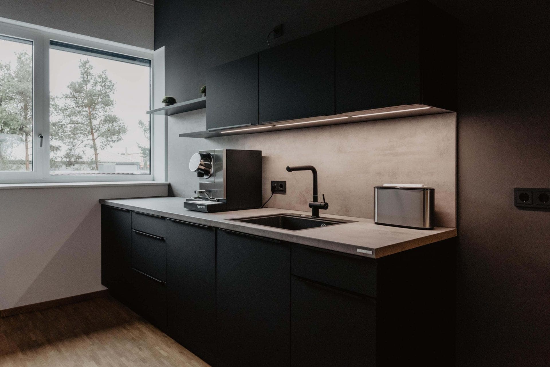 Kitchen counter with sink, coffee machine and concrete-look splashback beneath lit wall cabinets