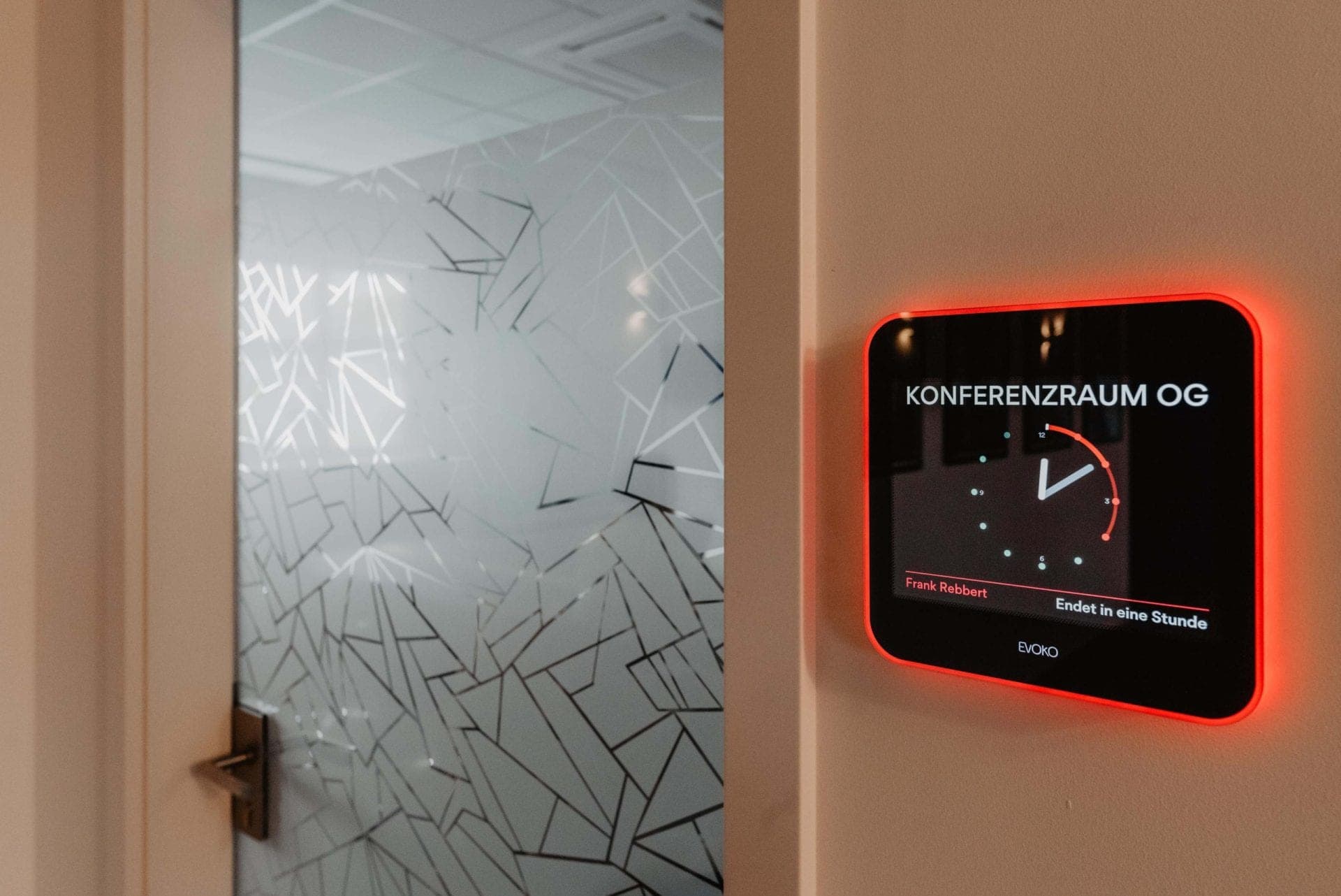 Digital room display reading Konferenzraum OG beside the glass door with a graphic pattern to the meeting room