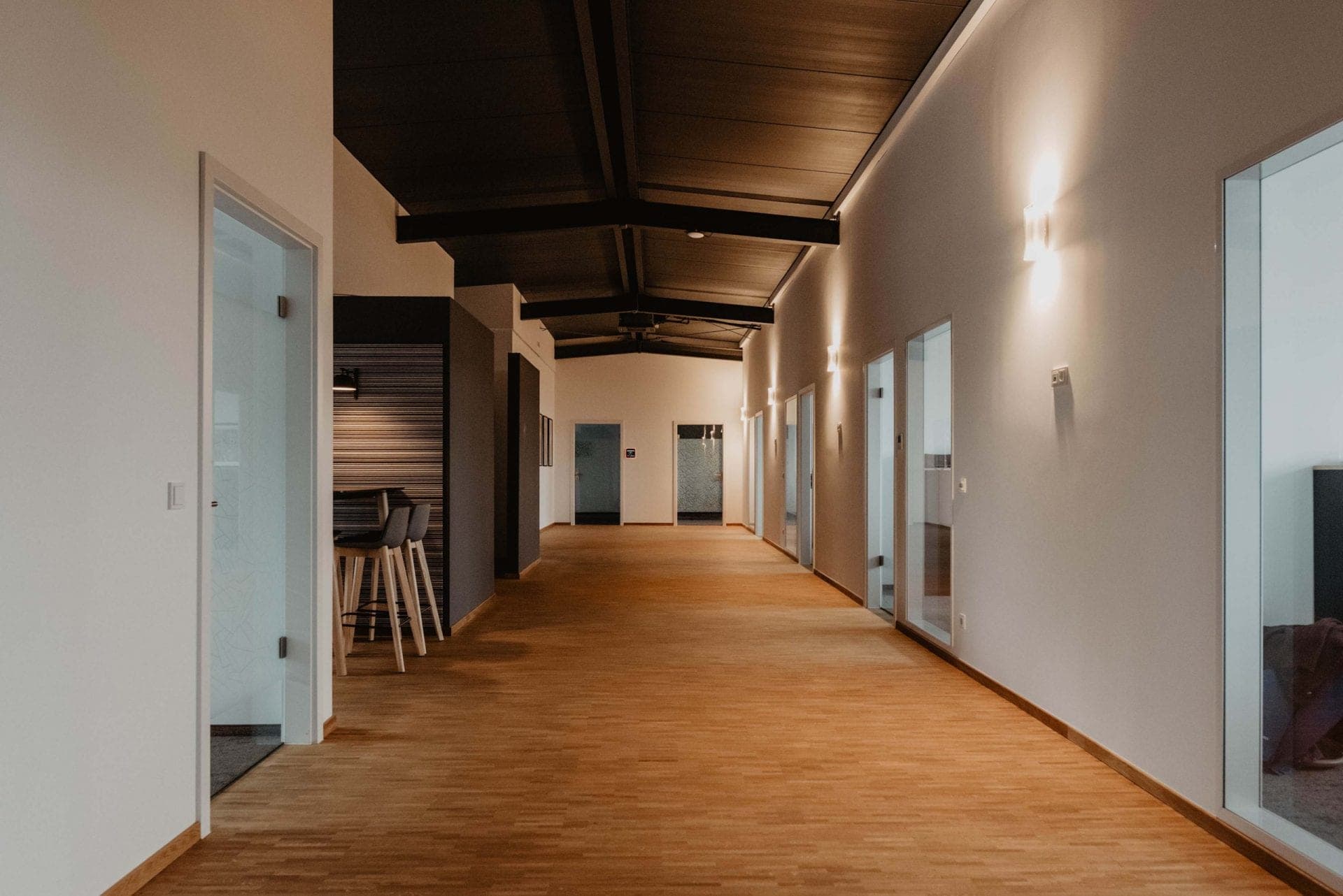 Further corridor with a dark beamed ceiling, parquet floor, glazed office doors and a standing table by the kitchenette