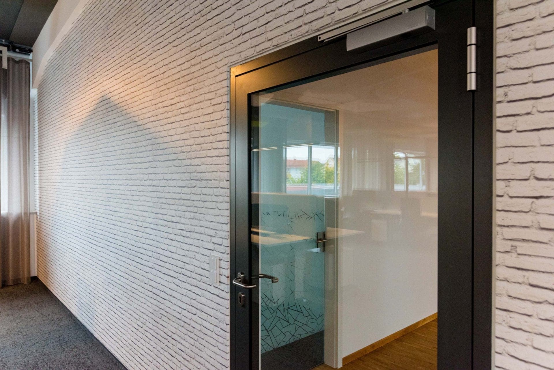 Corridor with a white-painted brick wall and an open dark-framed glass door leading to an adjoining room