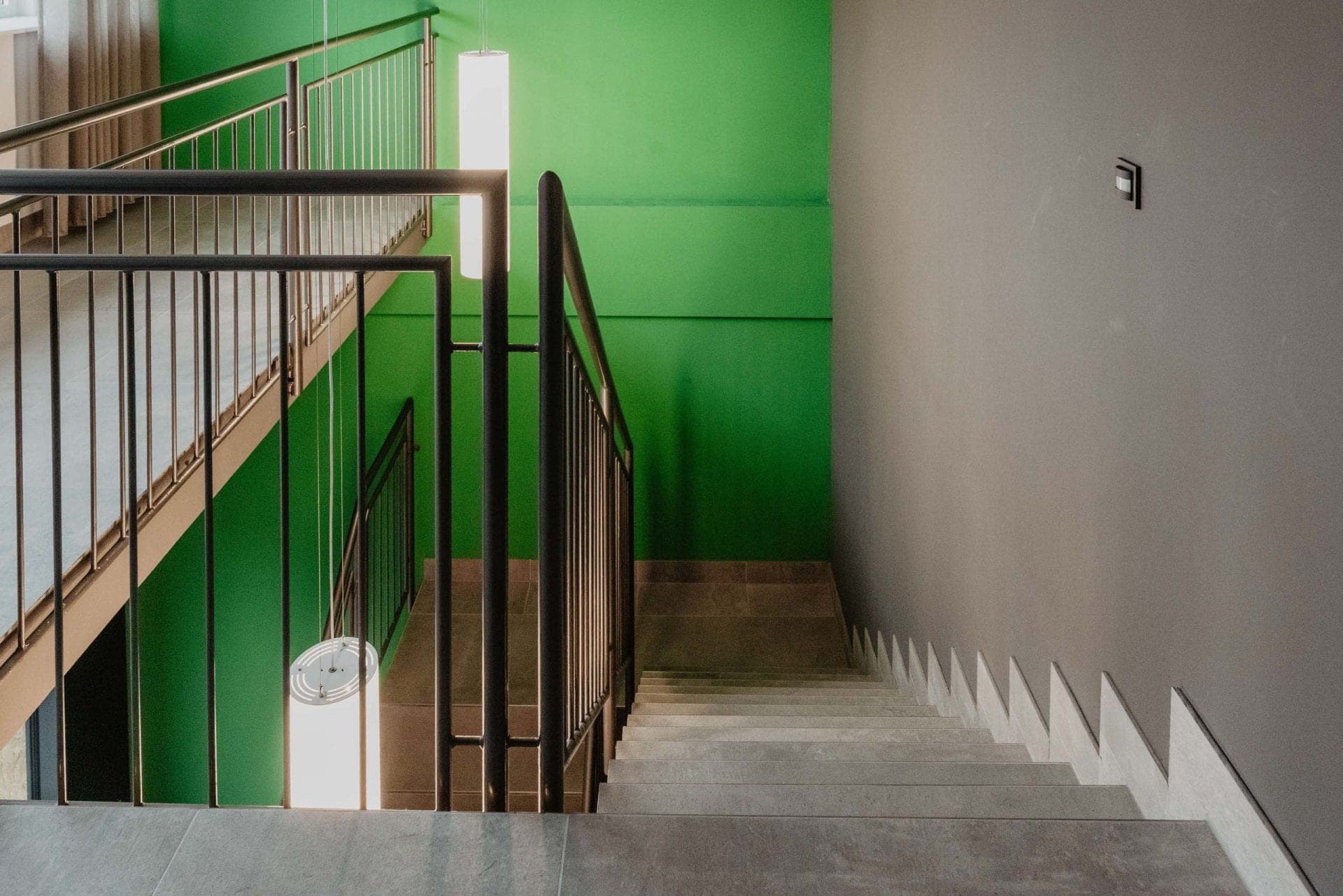 Staircase leading down past a green-painted wall, with a dark steel handrail and cylindrical pendant lights