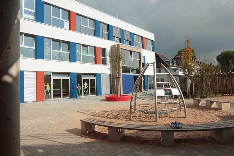 Schoolyard with a climbing arch in the sand, curved bench and tree seat in front of the glazed ground floor