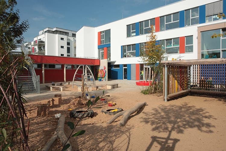 Play yard with logs, tyres and a climbing frame in front of a red single-storey building and the new school