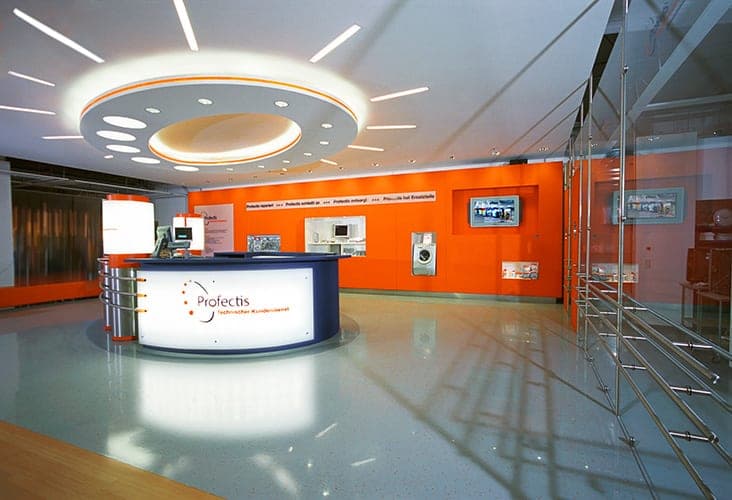 Round reception counter with the Profectis logo in front of an orange service wall with screens and display niches