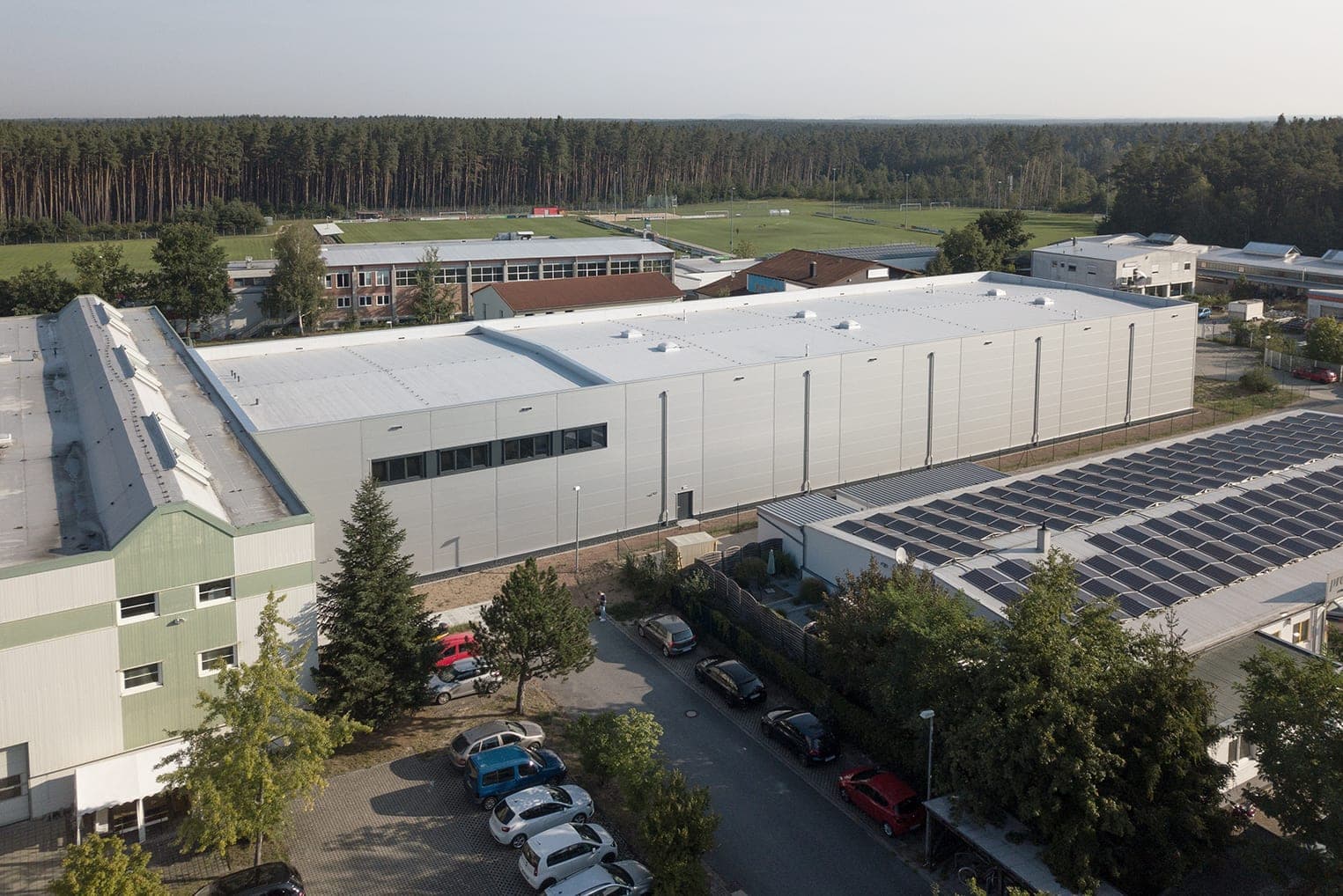Aerial view along the long side of the hall with existing buildings, sports pitches and forest in the background