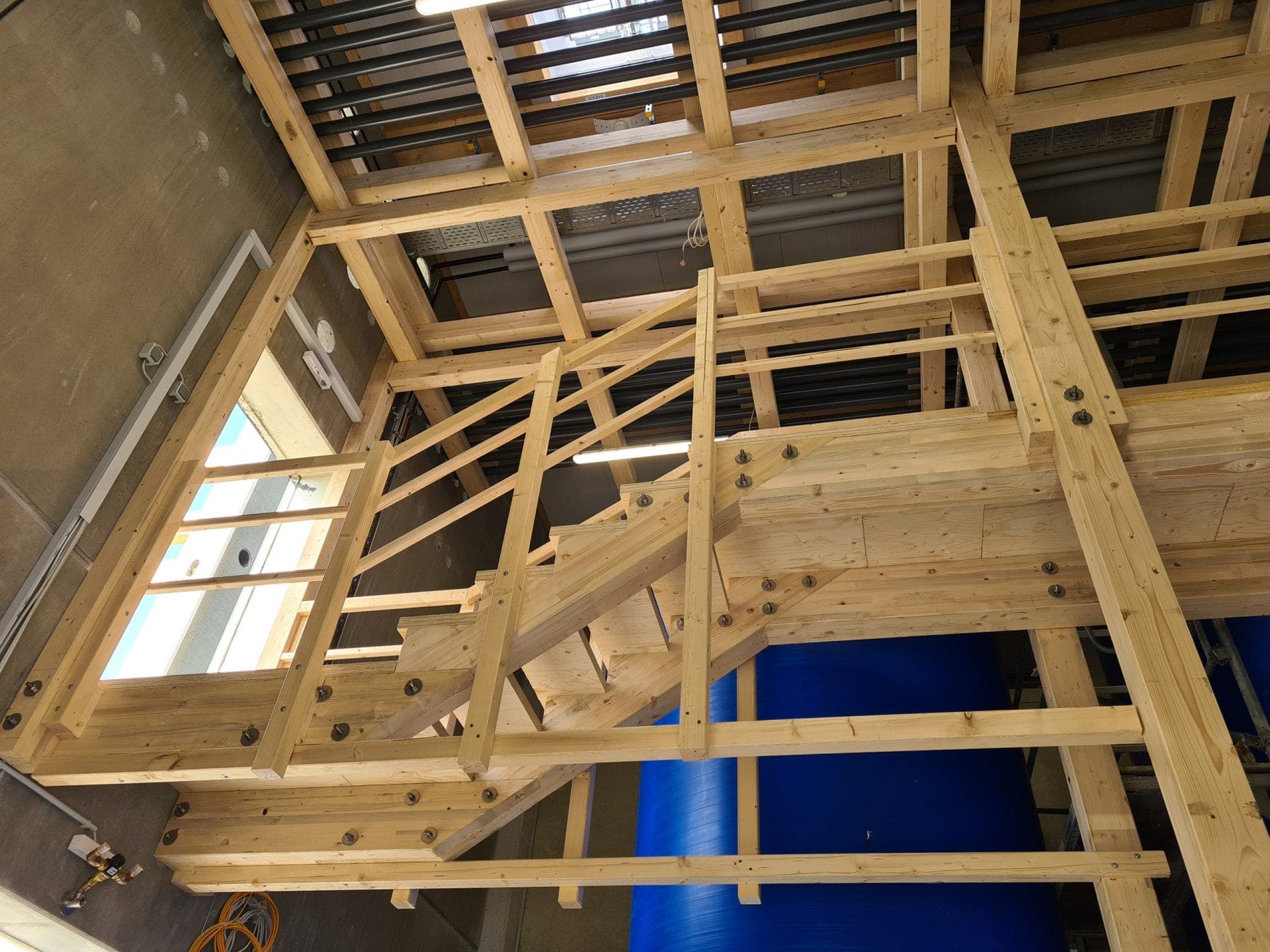 Holztreppe mit Podest und Geländer im Rohbau der Abfüllanlage, dahinter ein blauer Lagerbehälter