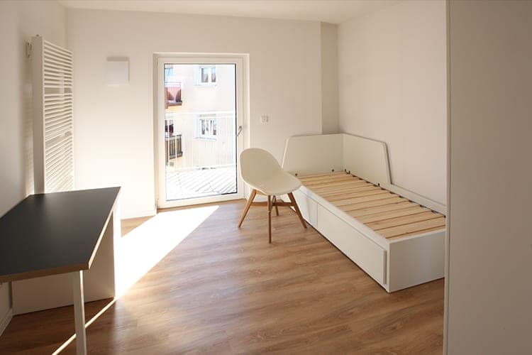 Empty student room with a bed, desk, wooden floor and a balcony door in sunlight