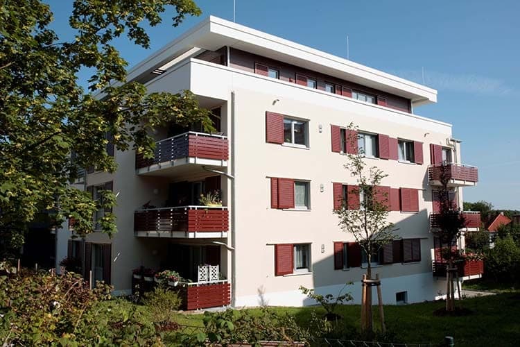 The residential building from the garden side with red balcony parapets, partly screened by a tree