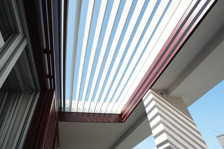 Upward view of the louvred canopy over a terrace, casting striped shadows on the column
